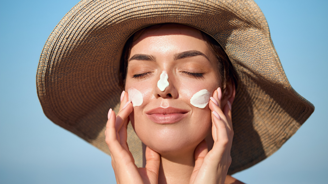 woman applying sunscreen to face wearing sun hat outdoors with blue sky, daily SPF skincare routine
